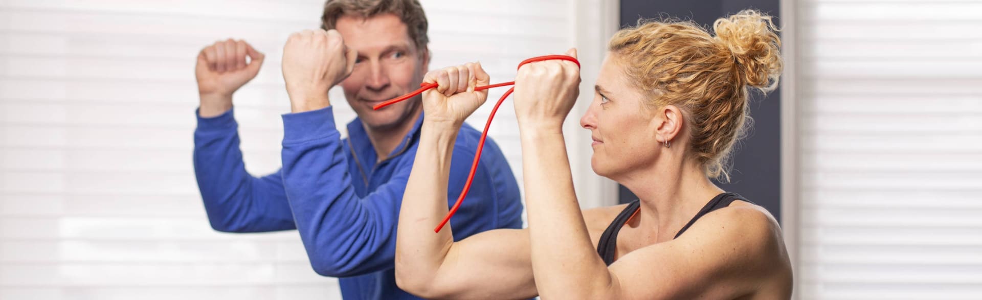 Een vrouw oefent met een rode weerstandsband, een fysiotherapeut doet de beweging voor.