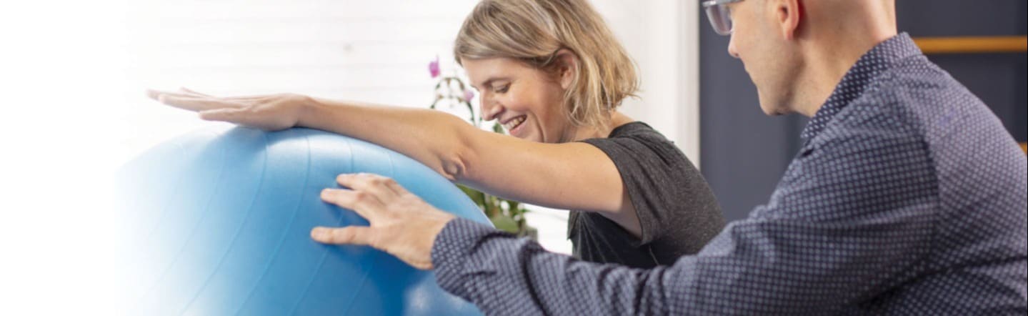 Een vrouw oefent lachend met haar armen op een grote blauwe bal, begeleid door een fysiotherapeut.